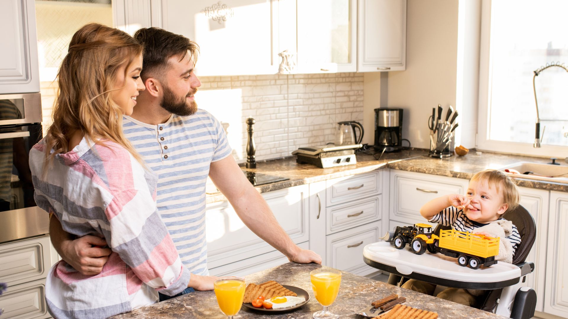 a man and a woman stand at a kitchen island with breakfast plates and orange juice. They smile at their infant, who is seated in a high chair, playing with a toy truck