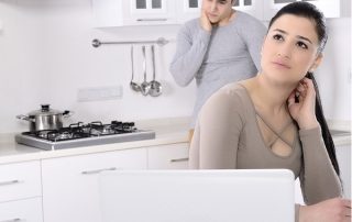 a man and a woman with worried expressions sit behind a laptop placed on a kitchen counter