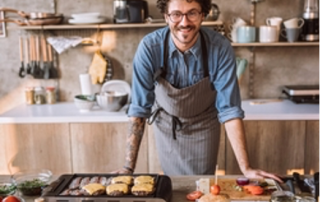 a man wearing an apron smiles and stands behind a countertop with food and plated dishes