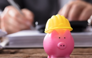 a small piggy-bank figurine wering a hard hat placed on a table where a person's hands are seen writing in the background